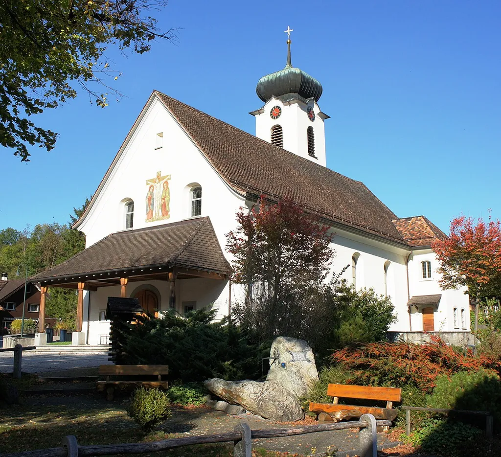 Süd- und Westfassade der Pfarrkirche Kobelwald. Links der überdachte Haupteingang. Rechts über dem Kirchendach erkennt man den Turm mit Zwiebeldach und Kreuz auf der Spitze. Die Turmuhr hat einen roten Grund, vergoldete Zeiger und römische Ziffern auf schwarzem Grund. Über dem Eingangsdach zwischen den zwei Emporen-Fenstern ist ein gemaltes Bild der Kreuzigungs-Gruppe. Vom gepflästerten Vorplatz führen drei Stufen zum Eingangsbereich, der mit vier auf Steinsockeln stehenden Holzsäulen markiert ist. Rechts an der Südostecke der Kirche ist die Sakristei angebaut. Im Vordergrund der Dorfpark mit Brunnen und Sitzbänken.