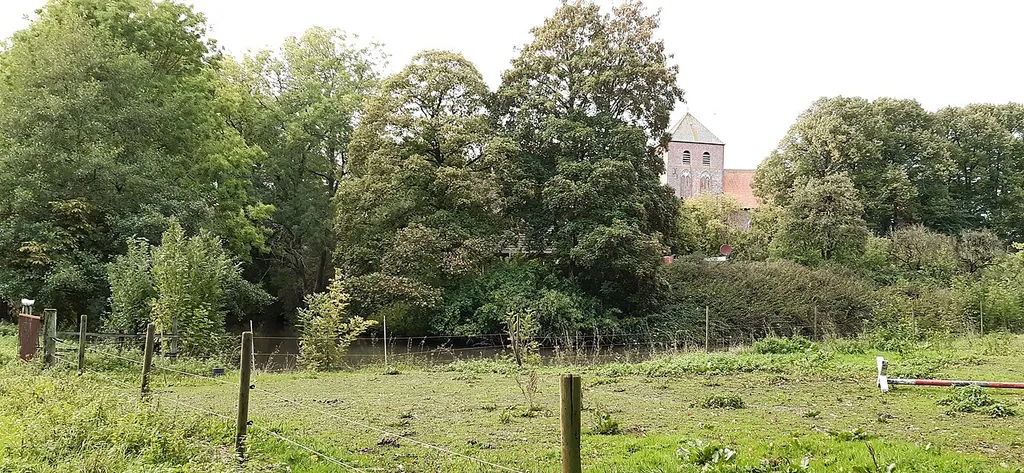 Blick auf den Burgstall Uttum von Süden; dahinter der schiefe Kirchturm der Uttumer Kirche, die nördlich direkt an den Wasserrgraben der Burg und des umgebenden Vorburgbereichs grenzte.