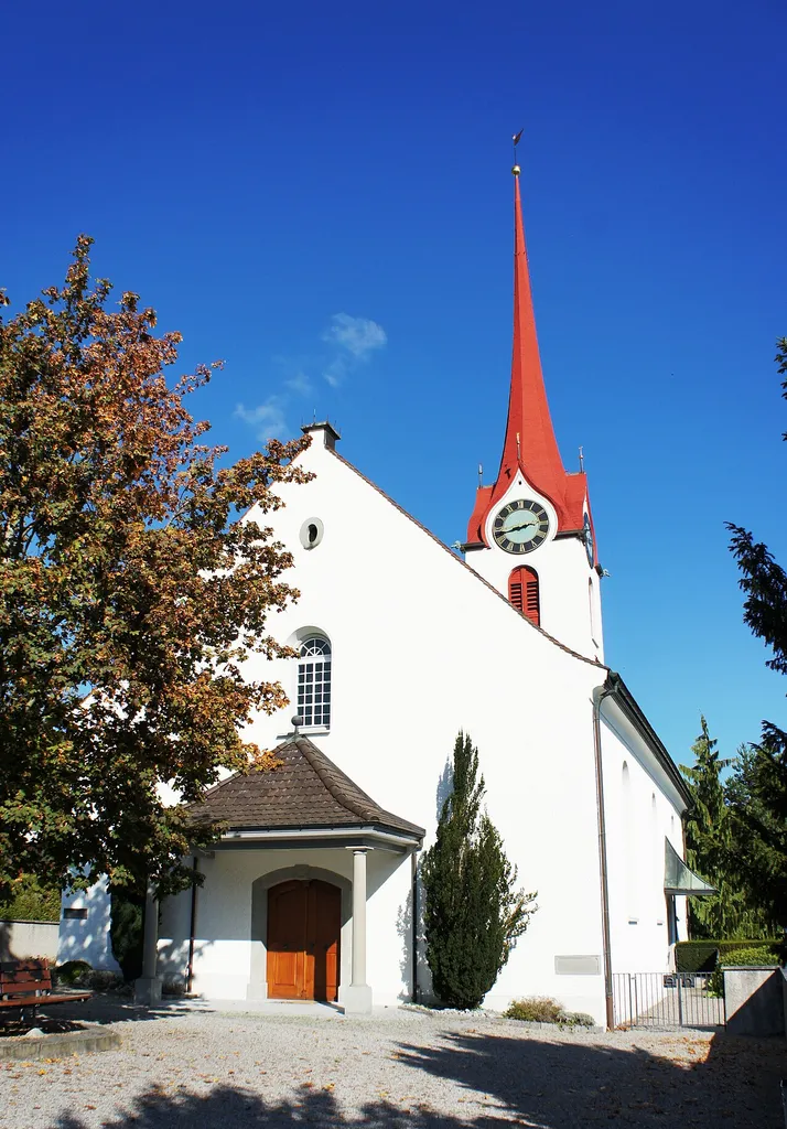 Die ref. Kirche in Rebstein SG aus Südwesten. Im Vordergrund der gepflästerte Vorplatz, in dessen Mitte steht ein Baum mit Sitzbänken umgeben. Stufenlos geht es zum überdachte Haupteingang an der Südwestfassade. Im Zentrum der Nordostfassade steht der Kirchturm mit Uhr und spitzigem, rotem Dach. Zuoberst ist eine Windfahne.
