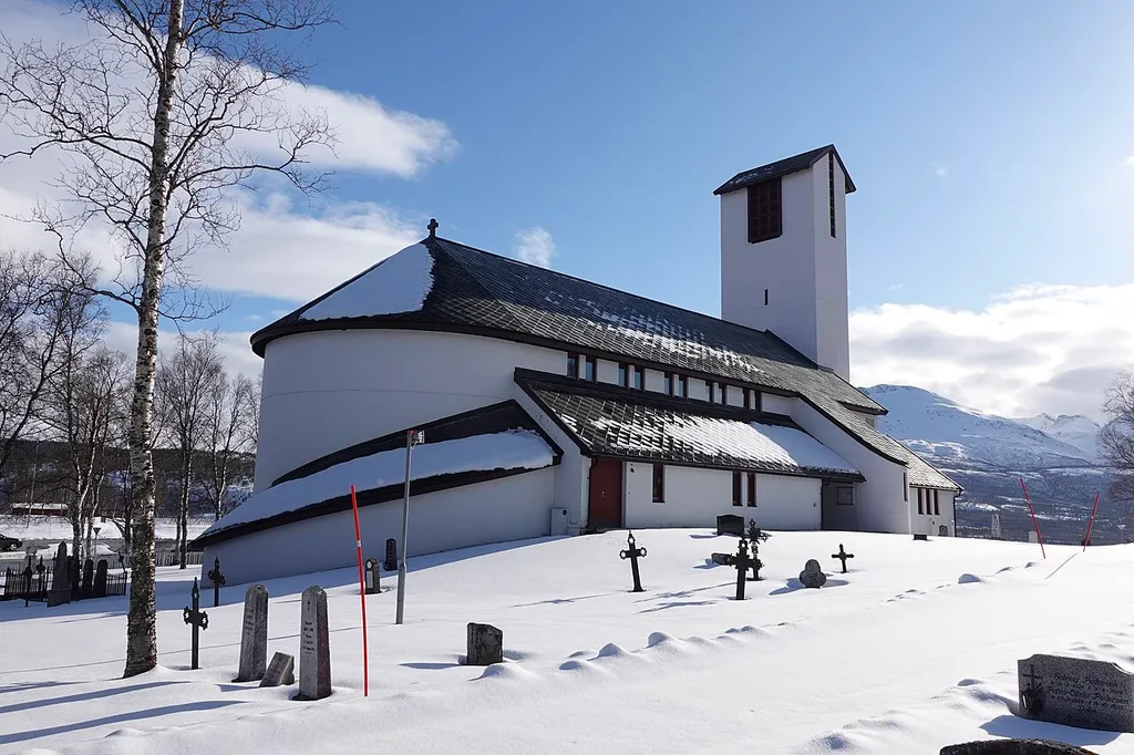 Salangen Church (Norwegian: Salangen kirke), a parish church of the Church of Norway in the village of Sjøvegan in Salangen Municipality in Troms og Finnmark county, Norway. The white concrete church with wood accents was built in a long church style in 1981 using plans drawn up by the architect Harald Hille. The church seats about 420 people. Cemetery/church yard with grave markers, etc. Photo taken on a spring evening in May 2022 with view towards the fjord and surrounding landscape, with the sun shining from a blue sky, and snow on the ground.
