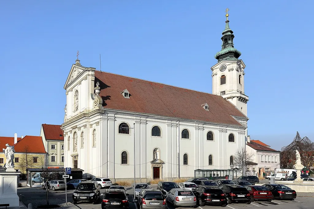 Südostansicht der röm.-kath. Pfarrkirche hl. Dreifaltigkeit in der niederösterreichischen Stadtgemeinde Bruck an der Leitha. Links der Sockel mit einer Statue der 1707 errichteten Mariensäule und rechts der 1713 erbaute Florianibrunnen.Die Kirche wurde von 1696 bis 1702 unter Einbeziehung des Stadtturmes errichtet. Dieser ist bis zum Schallgeschoss mittelalterlich (um 1230). Der Umlaufgang wurde im 16. Jahrhundert errichtet und das barocke Glockengeschoss um 1740. Die schmucklose Stirnfront wurde ebenfalls in dieser Zeit (1738–40) umgebaut. Unter anderem wurde sie mit einem geschosstrennenden Gebälk, einem giebelbekrönten Obergeschoss und einem dreiachsigen Mittelrisalit gegliedert. Zusätzlich wurden 6 Rundbogennischen für Steinstatuen eingebaut.