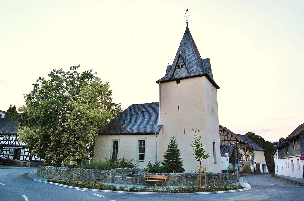 Kirche in Tiefenbach bei Braunfels mit mittelalterlichem (gotischem) Wehrturm (um 1300, Chorturm mit Schießscharten, Spitzhelm und vier Gauben) im alten Ortskern, umgeben von der alten Kirchhofmauer, die im Süden zu Gunsten des Straßenbaus versetzt wurde. Am Zugang von der Mittelstraße eine Kaiser-Wilhelm-Linde von 1913 (ist auf diesem Foto zu sehen - weiter links, am Rand dieses Fotos, von der Linde teilweise verdeckt, das älteste erhaltene Schulgebäude des Ortes, der Fachwerkbau wurde von 1684 bis 1847 als Schule genutzt, in den 1980/90er Jahren befand sich darin die Bäckerei Rudolf Kahl, wovon die gusseiserne Relieftafel und die goldfarbene Brezel an der Giebelseite zur Straße herrühren), an der Westwand der Kirche Kriegerdenkmal von 1924. Die Westhalle wurde 1559 ergänzt. Als der Bau 1713 bis 1714 um einen Nordflügel (Langhaus) zur Winkelhakenkirche erweitert wurde, erhielt das alte Schiff ein neues Portal, dessen aufwändiger Schmuck mit Inschrift und Wappen an den Bauherrn Graf Wilhelm Moritz zu Solms-Braunfels-Greifenstein und seine Frau Magdalena Sophie, geb. Prinzessin und Landgräfin zu Hessen-Homburg-Bingenheim erinnert. Im Okulusfenster über dem Portal die Initialen des damals betreuenden Kirchenbaumeisters Major Johann Georg Simon. Der winkelförmige Bau mit großen Fenstern und Walmdach wurde im Innern unter Aufgabe des alten Chores in Nordsüdrichtung umorientiert und neu ausgestattet: Kanzel datiert 1716, aber angeblich von 1687. Altar: einfacher Holztisch mit gedrechselten Beinen. Kirchenstuhl der Familie Simon mit Inschrift unter der L-förmigen Empore. Schönes Orgelprospekt mit reichen Schnitzereien, um 1715. Rankenmalereien an den Wänden aus der Umbauzeit (1713/14). (1980 wurden bei der Innenraumrenovierung die alten Ausschmückungen gefunden und freigelegt.) Glocke von 1601 durch Bernhard 1822 neu geschmiedet. Turmhahn von 1933.