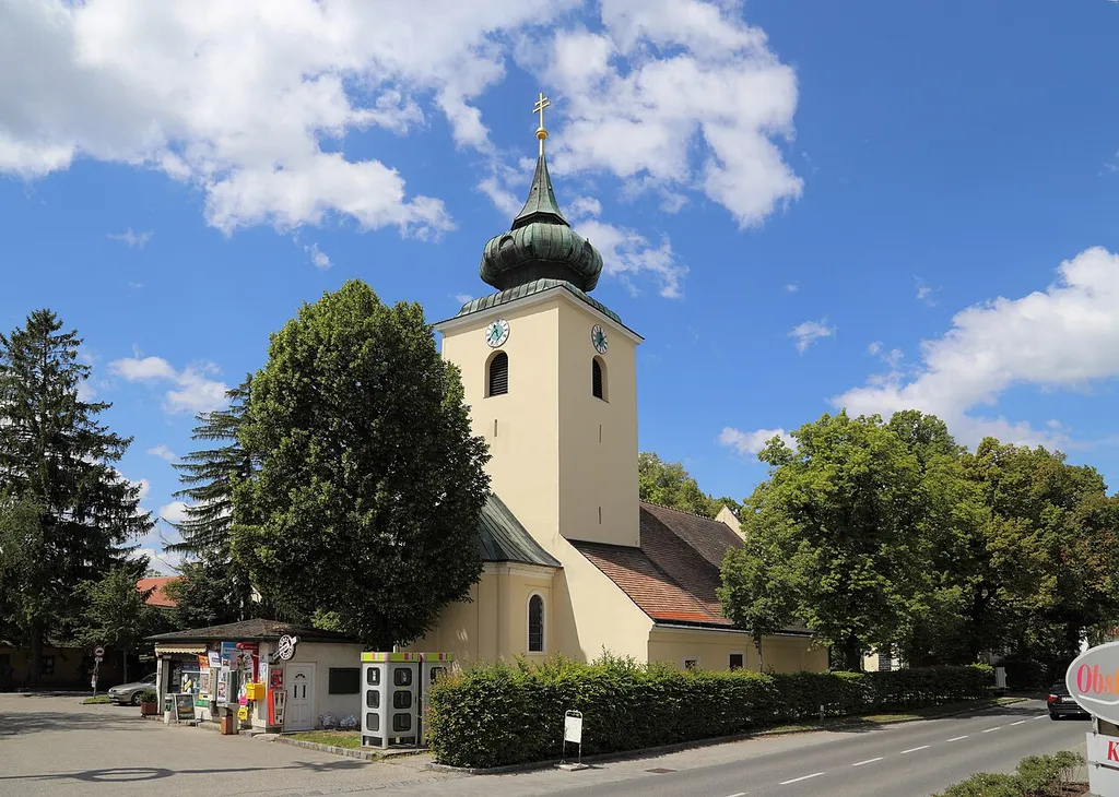 Nordostansicht der Pfarrkirche hl. Magdalena in der niederösterreichischen Gemeinde Raasdorf. Ein schlichter Sakralbau in der Mitte des Angers. Der gedrungene Ostturm (mit einer Biedermeierhaube) wurde in der 2. Hälfte des 13. Jahrhunderts über einem romanischen Chorquadrat errichtet, das Langhaus und der Chor mit rundem Schluß im Jahr 1736.
