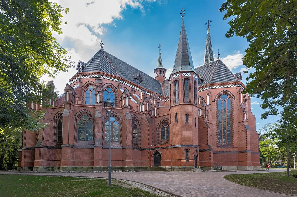 Cathedral of Sts. Peter and Paul in Gliwice, Gliwice, Silesian Voivodeship, Poland, on 15 September 2024. The photograph shows the Gothic Revival brick body of the cathedral as seen from the surrounding greenery. The frame includes brick elevations of the church, pointed-arch windows, buttresses, decorative brick details, tiled roofs, slender turrets and tall spires, as well as a paved path, a park lamp, trees and greenery around the church. The image documents the exterior architecture of the cathedral, its Gothic Revival character, the surroundings of the church and public space in central Gliwice.
