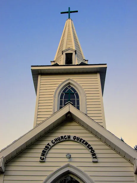This photo depicts the spire of the Christ Church in Queensport. Nova Scotia, Canada. There is a cross on top of the spire. There is an arched window. the top of the door is visible.