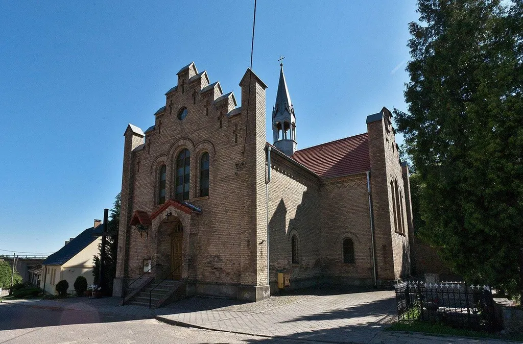 Trudny do sfocenia, za obsadzony wysoką zielenią neogotycki kościół z poł. XIX w. Na przeciwko, po drugiej stronie ulicy, budynek plebanii, dziś wykorzystywany jako dom mieszkalny.
