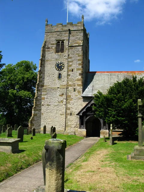 West tower of All Saints' parish church, Pickhill, North Yorkshire, seen from the south. In the foreground is a sundial set on top of the broken shaft if a medieval churchyard cross.