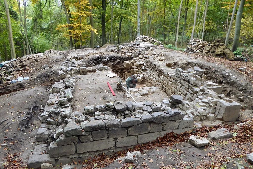 Archäologische Grabung an einer Kirchenruine nahe Asche, Stadt Hardegsen, Südniedersachsen. Die mittelalterliche Kirche war innenseitig verputzt, sie wies einen Turm (ganz hinten), ein eingewölbtes zweijochiges Kirchenschiff und einen Chorraum (vorne) auf, der wahrscheinlich mindestens einmal erweitert wurde. Der Ostteil des Chorraums war gegenüber dem restlichen Teil noch einmal erhöht, das originale Fußbodenniveau konnte festgestellt werden (nördlicher, im Bild rechter Bereich des Chorraums). Die Außenwand wies eine Sockelabschrägung aus sorgfältig behauenen, geflächten Steinen auf. Im Süden des Chorraums befand sich ein Zugang für den Priester.