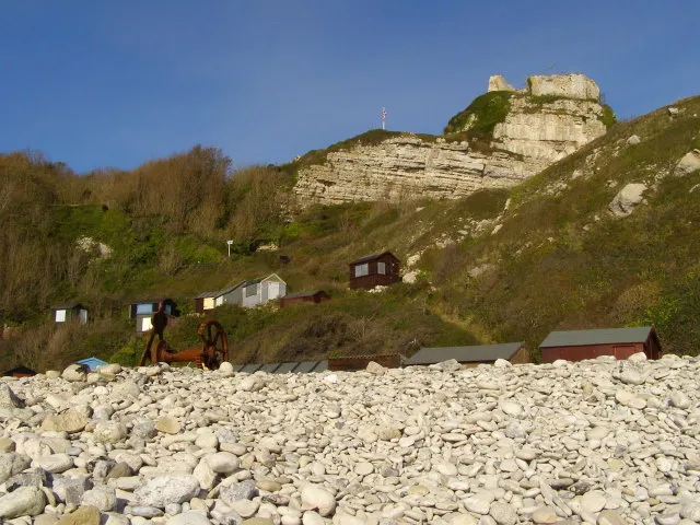 Church Ope Cove Looking up the bank of the pebble beach, with the beach huts and castle ruins beyond. At the top of the pebble bank, on the left, are the rusting remains of a boat winch.