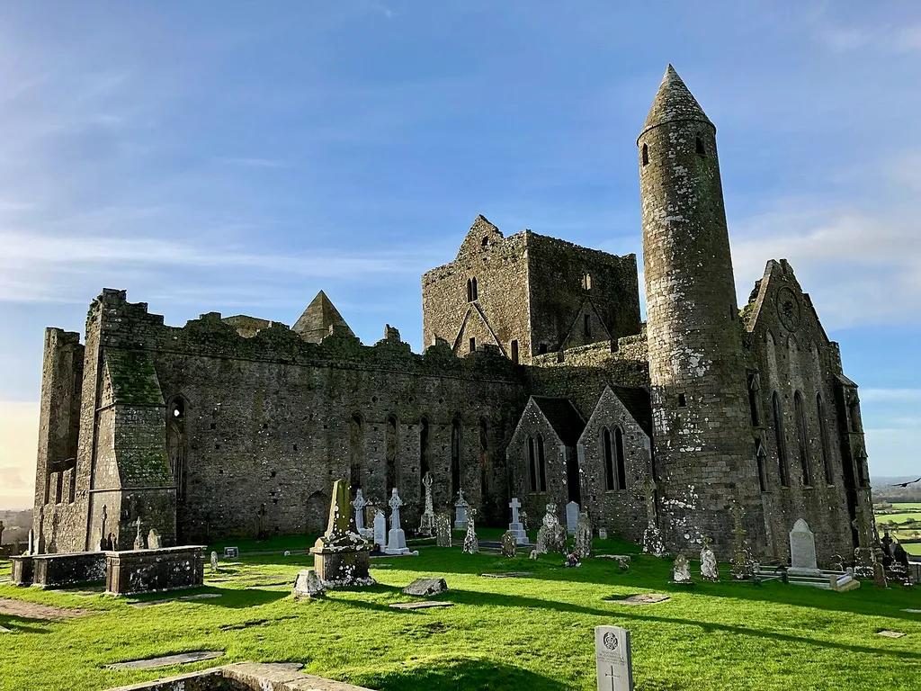 This is the Carraig Phádraig, or the Rock of Cashel, also known as St. Patrick’s Rock, located in the town of Cashel in County Tipperary, Éire. Originally the site of a hilltop fort, home to the kings of Munster for many centuries, the Rock was donated to the church in 1101 by King Muirchertach Ua Briain. However, few 12th Century structures survive, with the round tower, 90 feet (28 meters) tall, and Cormac’s Chapel, a Romanesque-style sandstone Building, was constructed atop the rock between 1127 and 1134, being the only buildings left from this time period. The largest of the structures atop the rock is The Cathedral, which was built between 1235 and 1270, which abuts against a residential structure for the clergy, known as the castle, constructed in the 15th Century. Both of these buildings are now in ruins. The other building on the site is the Vicars Choral, constructed in the 15th Century, which today has been fully restored, and houses several Celtic and medieval artworks. The rock remained intact and in use for major religious functions until 1647, when it was sacked during the Irish Confederate Wars, after Irish Confederate troops sought refuge on the rock, and were massacred by the opposing English Parliamentarian troops, along with the Catholic clergy, whom the English Parliamentarian troops saw as aiding the enemy. The buildings were sacked and vandalized. The buildings remained intact, however, though in a state of partial ruin, until 1749, when the main cathedral roof was removed by the Anglican Archbishop of Cashel, showing how little regard or respect the English had for the Irish people. The rock became home to a cemetery during the 18th and 19th Centuries, with many impressive grave markers. The most notable feature of this is a large tomb built for the Scully Family in 1867, once topped by a large cross, destroyed in 1976 by lighting, which sits in pieces on the ground nearby. The buildings on the rock have had conservation work carried out on th