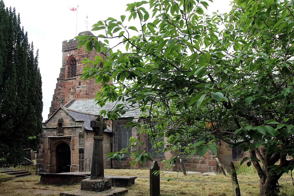 View of Grade II* listed Holy Cross church in Woodchurch, Birkenhead, showing buttressed tower and porch. In front is a standing cross, a Scheduled Ancient Monument.