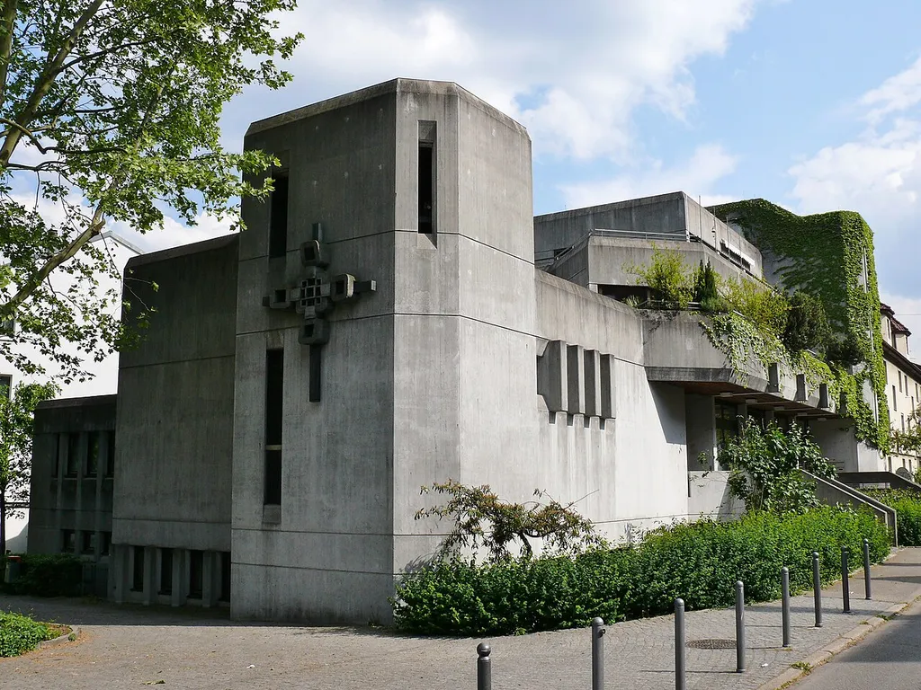 Die Heilig Geist Kirche in Stuttgart-Ost. The raw concrete structure was built 1974-1976 to plans by Rainer L. Neusch. It is a community center with integrated church. The church space can be enlarged by opening a folding wall with the community hall. Altar design and bronze work (portals, cross, tabernacle) by the sculptor Gerhard Tagwerker. The outer cross, the abstract Way of the Cross and windows were created by Gudrun Müsse-Florin. Late Gothic Madonna around 1480. Previous buildings from 1930 (previously Notkirche of St. George, destroyed 1944) and 1946/47. Ref: https://de.wikipedia.org/wiki/Liste_der_Kirchen_in_Stuttgart#Römisch-katholische_Kirchen