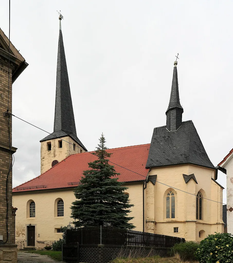 Die Kirche ist im Kern romanisch und der Turm eine gotische Zugabe, das fällt von außen kaum ins Auge. Der Chor wirkt allerdings auch von außen sofort gotisch. Der Turmhelm und seine Schrägstellung sind von weitem zu sehen, doch liegt diese nicht an strukturellen Schäden, sondern man hat ihn bewusst gegen die Hauptwindrichtung geneigt. Die Chorapsis weicht architektonisch auffällig ab, möglicherweise sollte sie der Anfang eines Neubaues werden, der dann aufgegeben werden musste. Das links angeschnittene Gebäude ist die ehemalige Dorfschule.