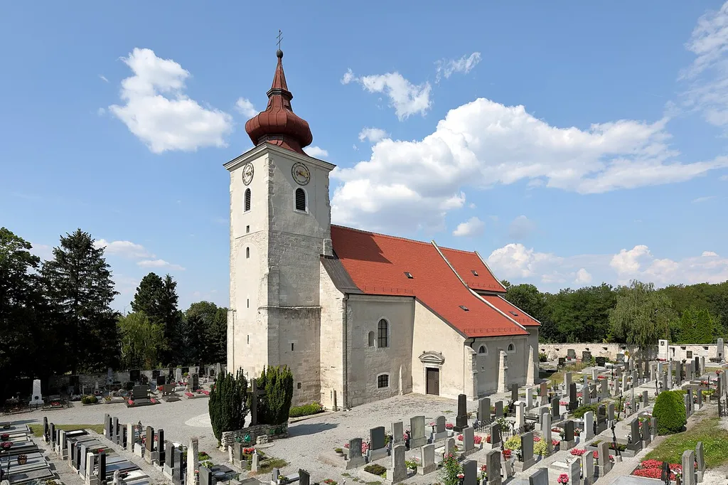 Südansicht der Pfarrkirche hl. Petronella in der niederösterreichischen Marktgemeinde Petronell-Carnuntum. Eine zu Beginn des 13. Jahrhunderts errichtete romanische Kirche, die teilweise von einer mittelalterlichen bewehrten Friedhofsmauer umgeben ist. Die Errichtung des Glockengeschosses (inklusive Rundbogenfernstern, Turmuhr und Zwiebelhelm) des Kirchturmes sowie die Erhöhung des Langhauses erfolgte Ende des 17. Jahrhundert. Die südliche Seitenkapelle stammt aus dem Ende des 14. Jahrhunderts.