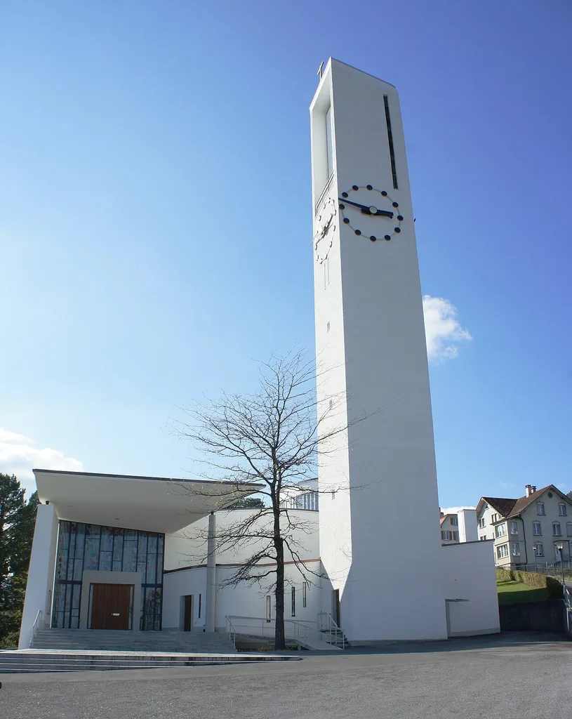 Die kath. Kirche Rebstein aus Osten. Links zum überdachten Haupteingang führen sowohl Stufen als auch eine rollstuhlgängige Rampe. Im mittleren Bildteil steht der Kirchturm mit Turmuhren und Aussichtsplattform auf Höhe des Glockenstuhls. Rechts vom Turm ist die Sakristei. Am rechten Bildrand ist der Burgweg, der zum Friedhof hinauf führt, zu erkennen.