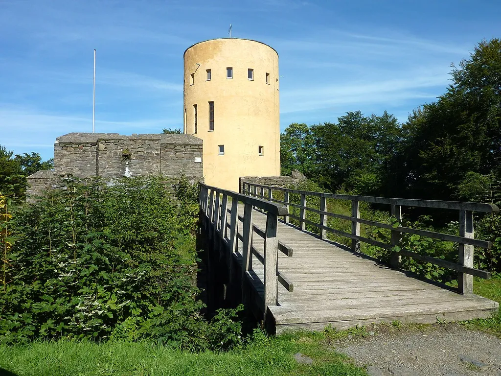 Gemeinde Grund (Hilchenbach)/Westfalen: Die Ginsburg-Ruine, vom 13. bis 17. Jahrhundert Grenzfestung des Hauses Nassau auf dem Schlossberg, Ansicht von Südwesten. Links im Bild ein Teil des rekonstruierten äußeren Mauerrings. In der Bildmitte der in den Jahren 1967/68 erbaute Aussichtsturm, den ehemaligen Bergfried nachahmend und auf dessen etwa aus dem Jahr 1200 stammenden Fundamenten errichtet. Im Vordergrund eine in der Gegenwart errichtete Holzbrücke über den Burggraben.