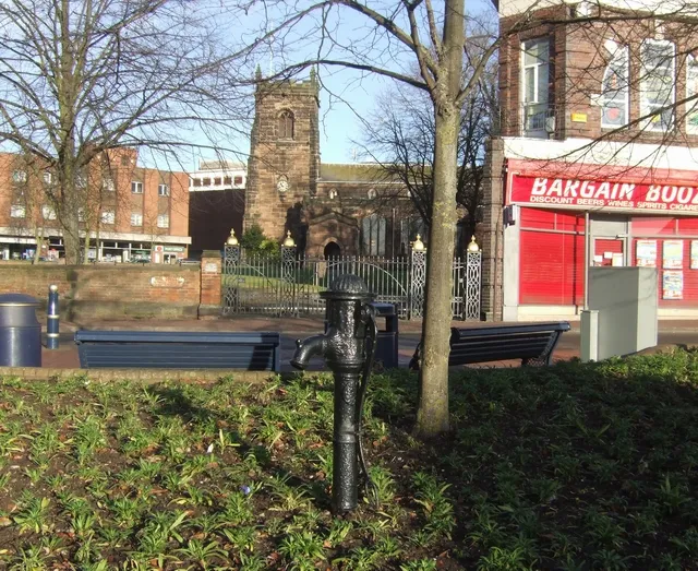 Cast iron pump in Mill Street, Cannock, Staffordshire, with St Luke's parish church in the background. The Georgian iron gates to the churchyard are Greade II listed.
