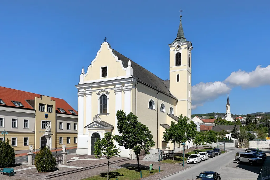 Südostansicht der röm.-kath. Pfarrkirche hl. Katharina am Hauptplatz in der burgenländischen Marktgemeinde Rechnitz und links das Gemeindeamt sowie rechts hinten die evangelische Pfarrkirche.